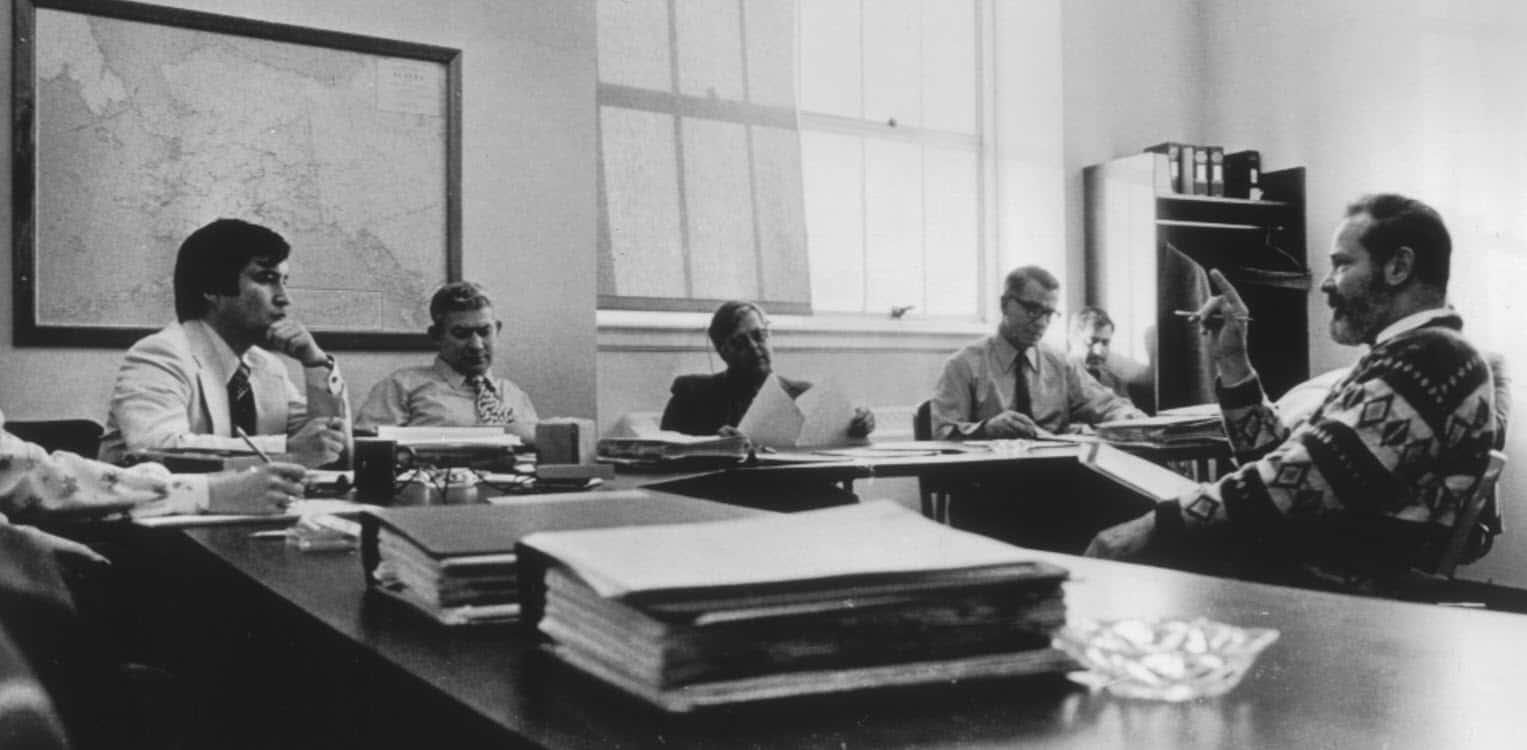 5 men sitting around a table with paperwork
